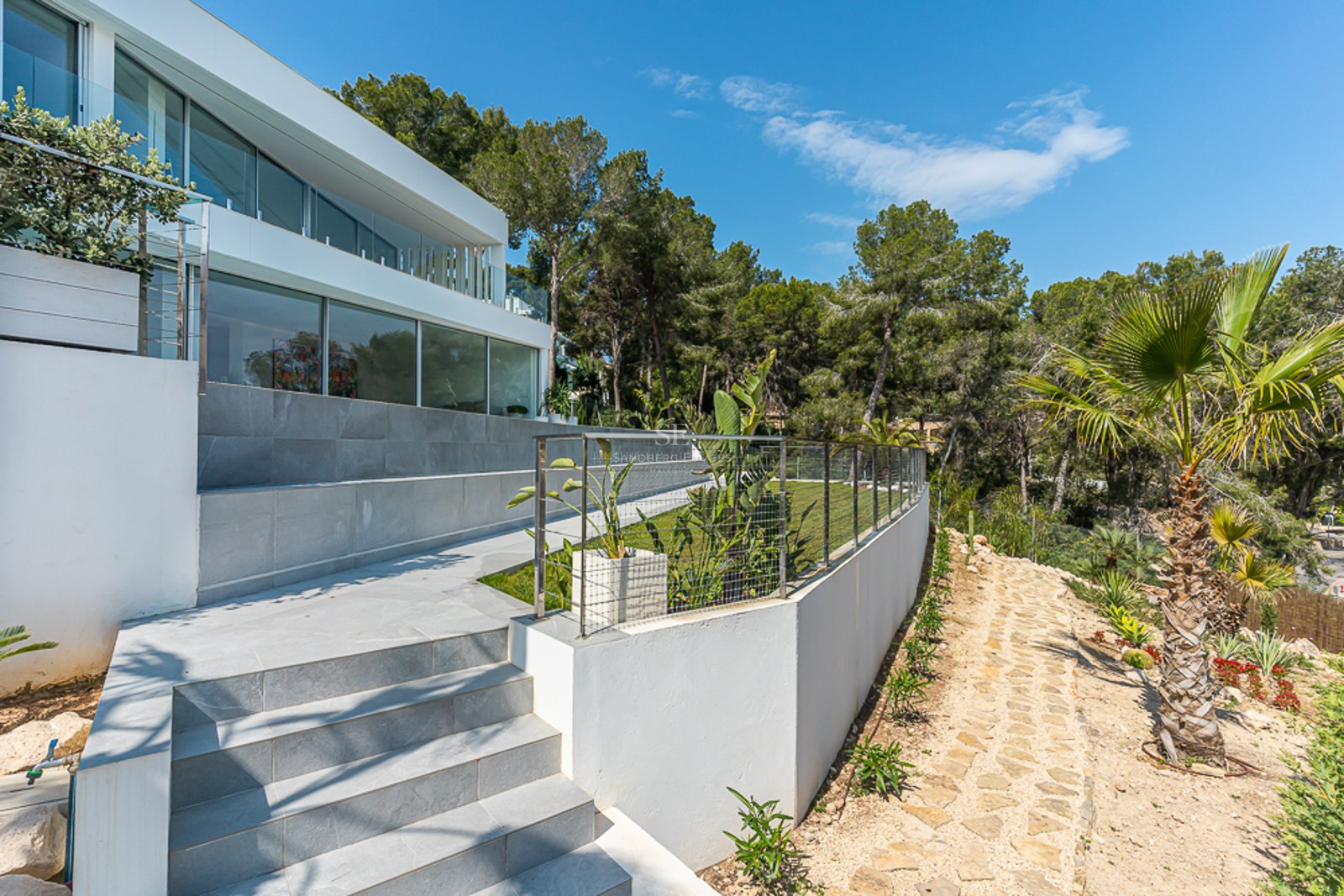 Modern white villa with a tiered garden featuring stone stairs, a lawn, and palm trees under a clear blue sky.