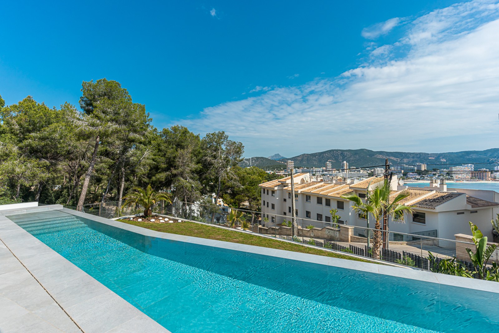 Luxury infinity swimming pool with stone tiling, overlooking a coastal town, mountains, and the sea under a clear blue sky.