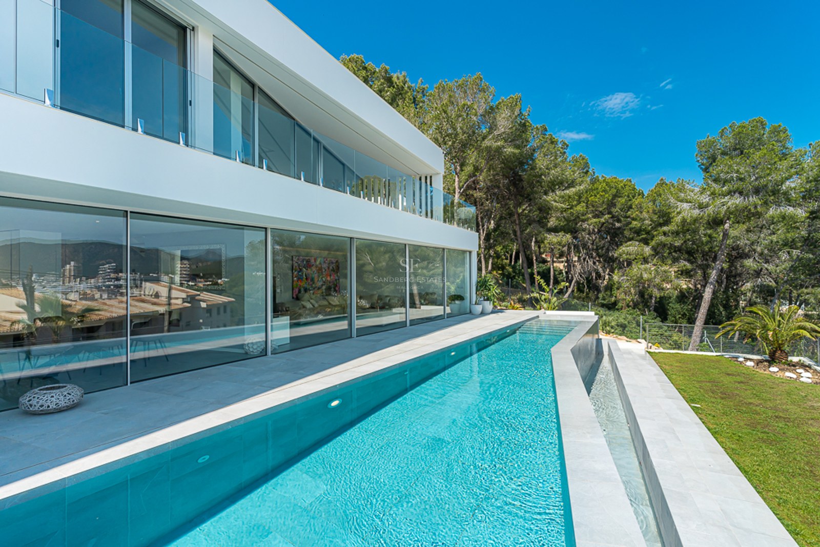 A sleek modern white villa with a long turquoise lap pool, floor-to-ceiling glass windows, and lush green trees.
