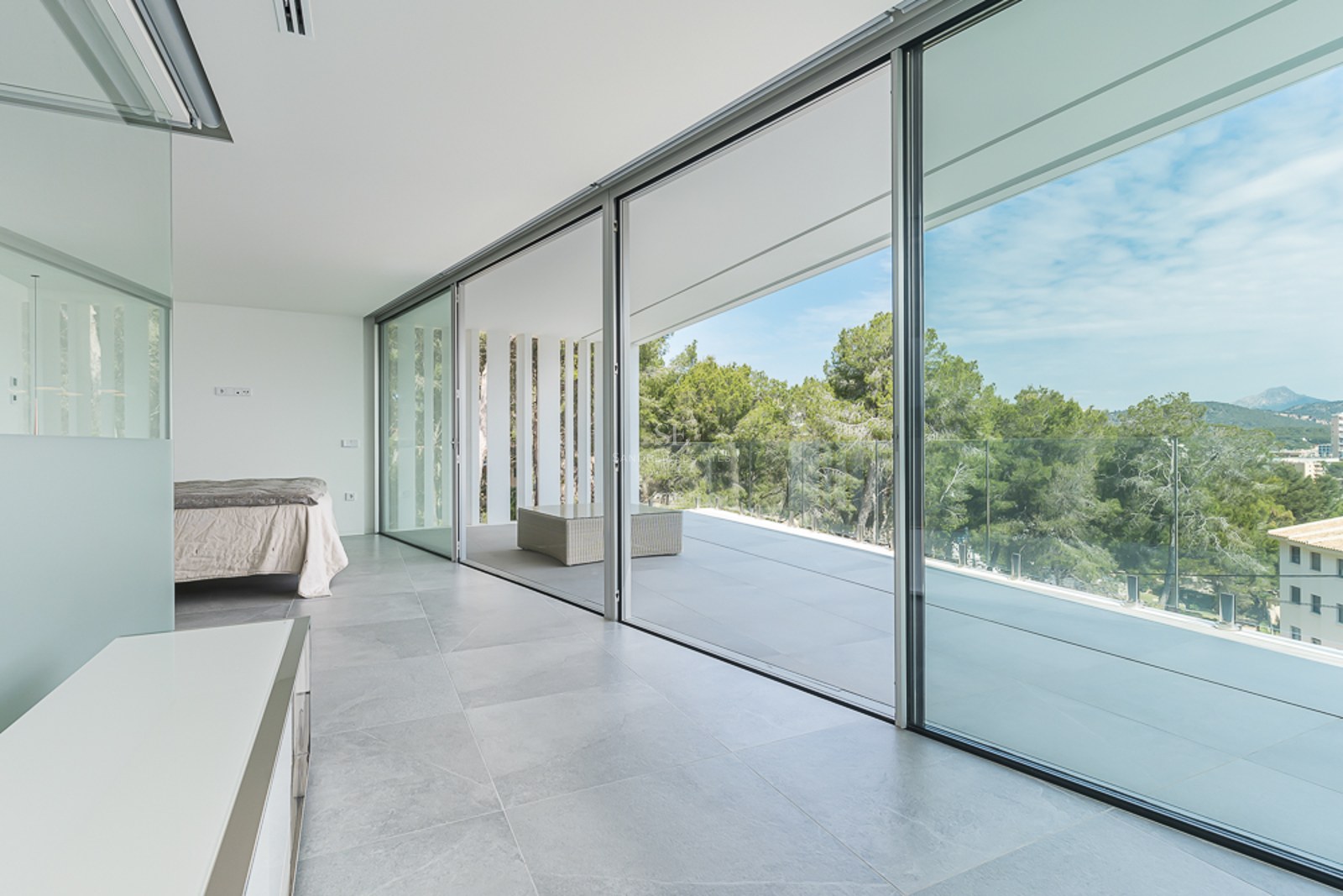Bright minimalist bedroom with large sliding glass doors opening to a terrace with forest views.