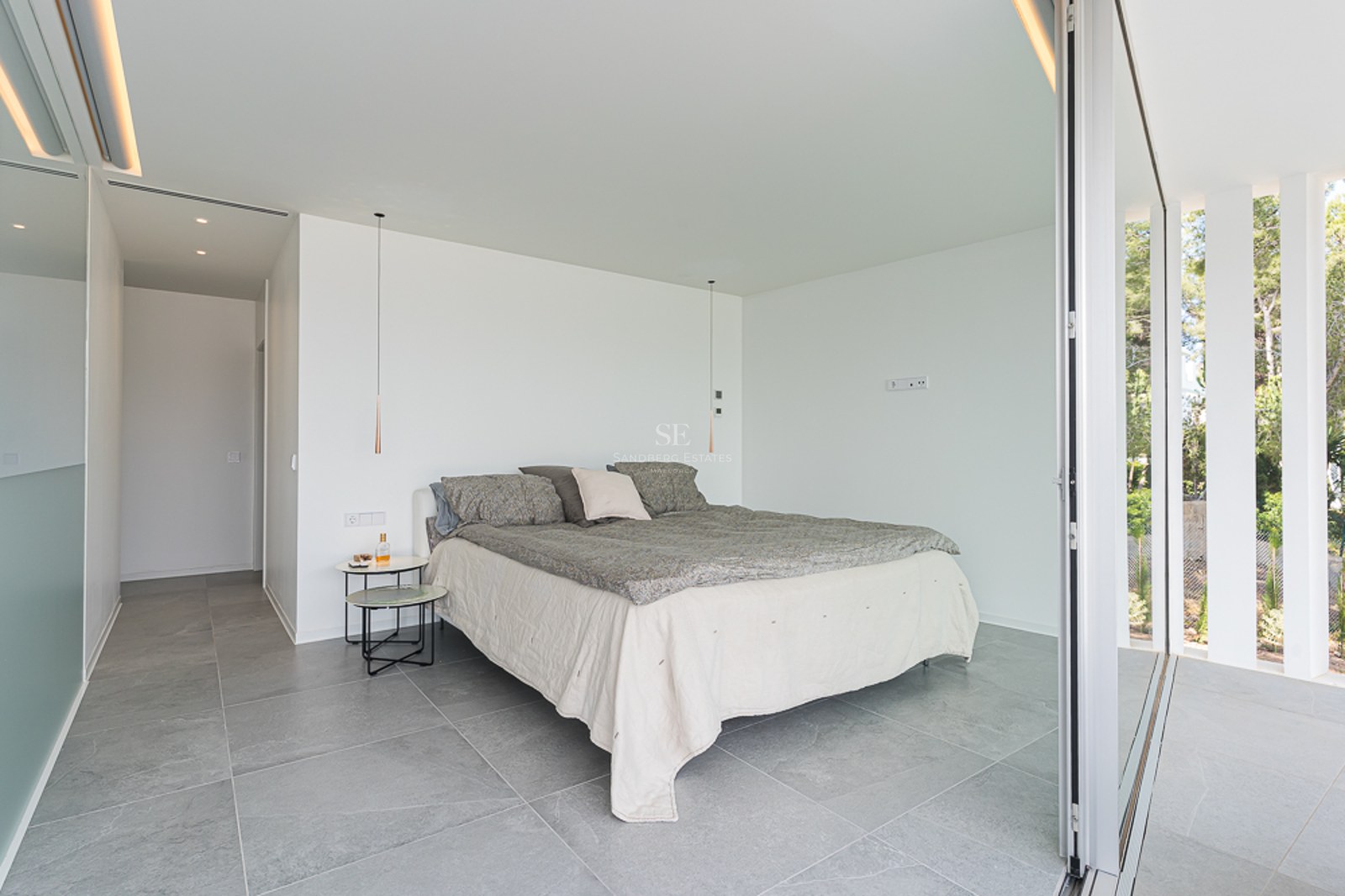 Minimalist bedroom with grey stone floors, white walls, and a large sliding glass door opening to a garden.