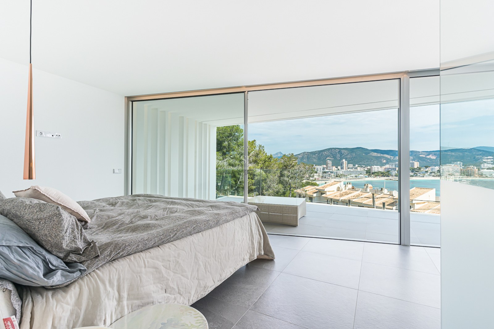 Bright modern bedroom with a large bed and floor-to-ceiling glass doors opening to a terrace with coastal views.