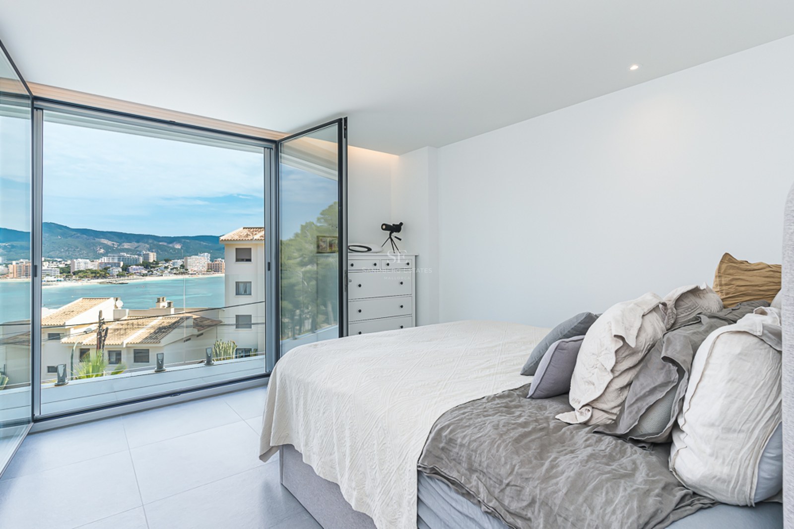 Bright bedroom with a bed and large glass doors opening to a coastal bay view and mountains.