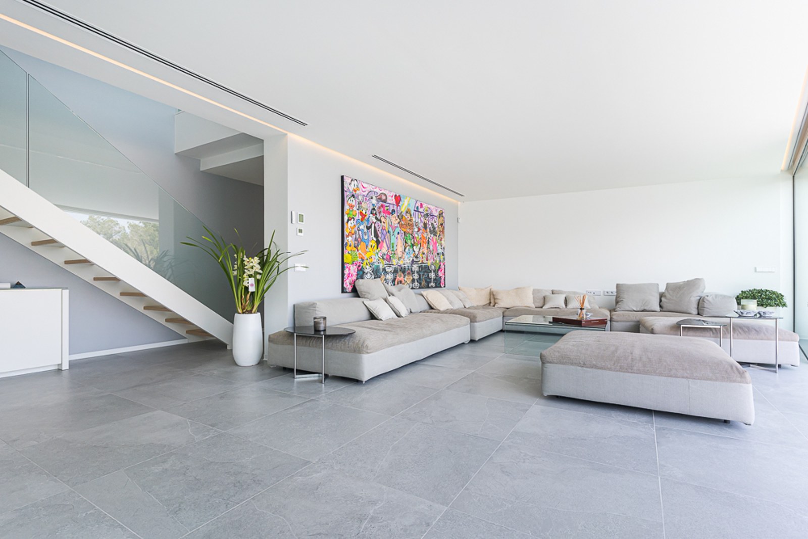 Spacious living room with large grey modular sofa, colorful wall art, and glass-railing staircase.