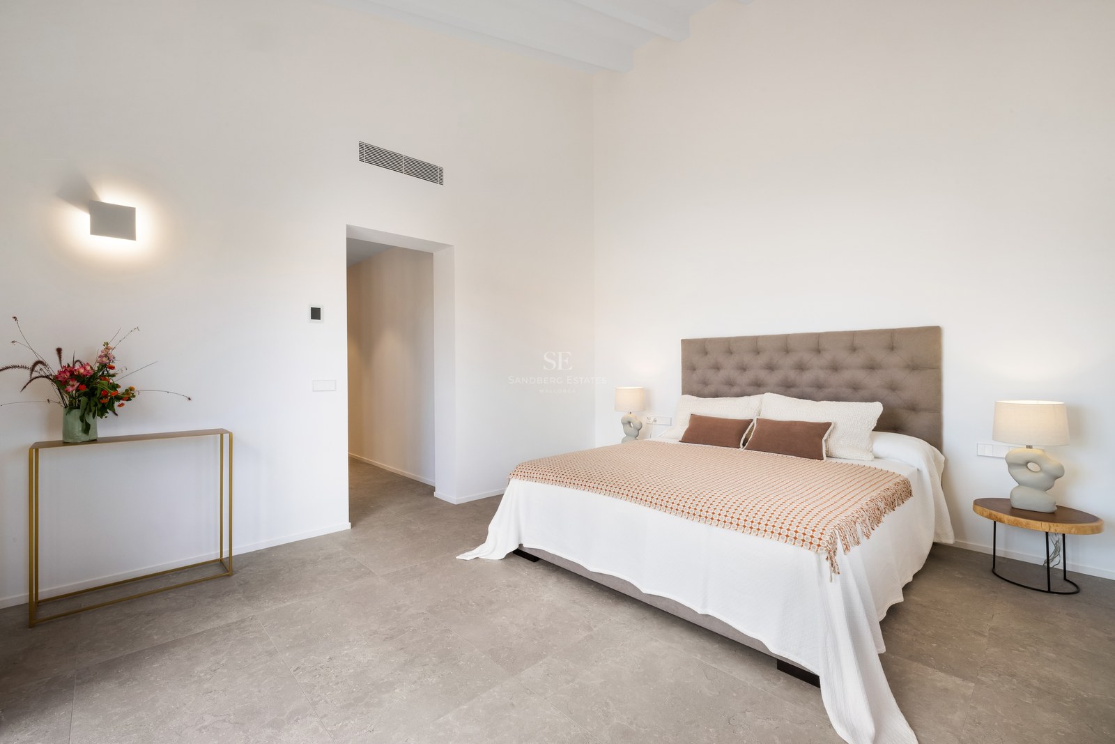 Minimalist bedroom with a tufted headboard, white bedding, designer lamps, and grey stone floors under high ceilings.