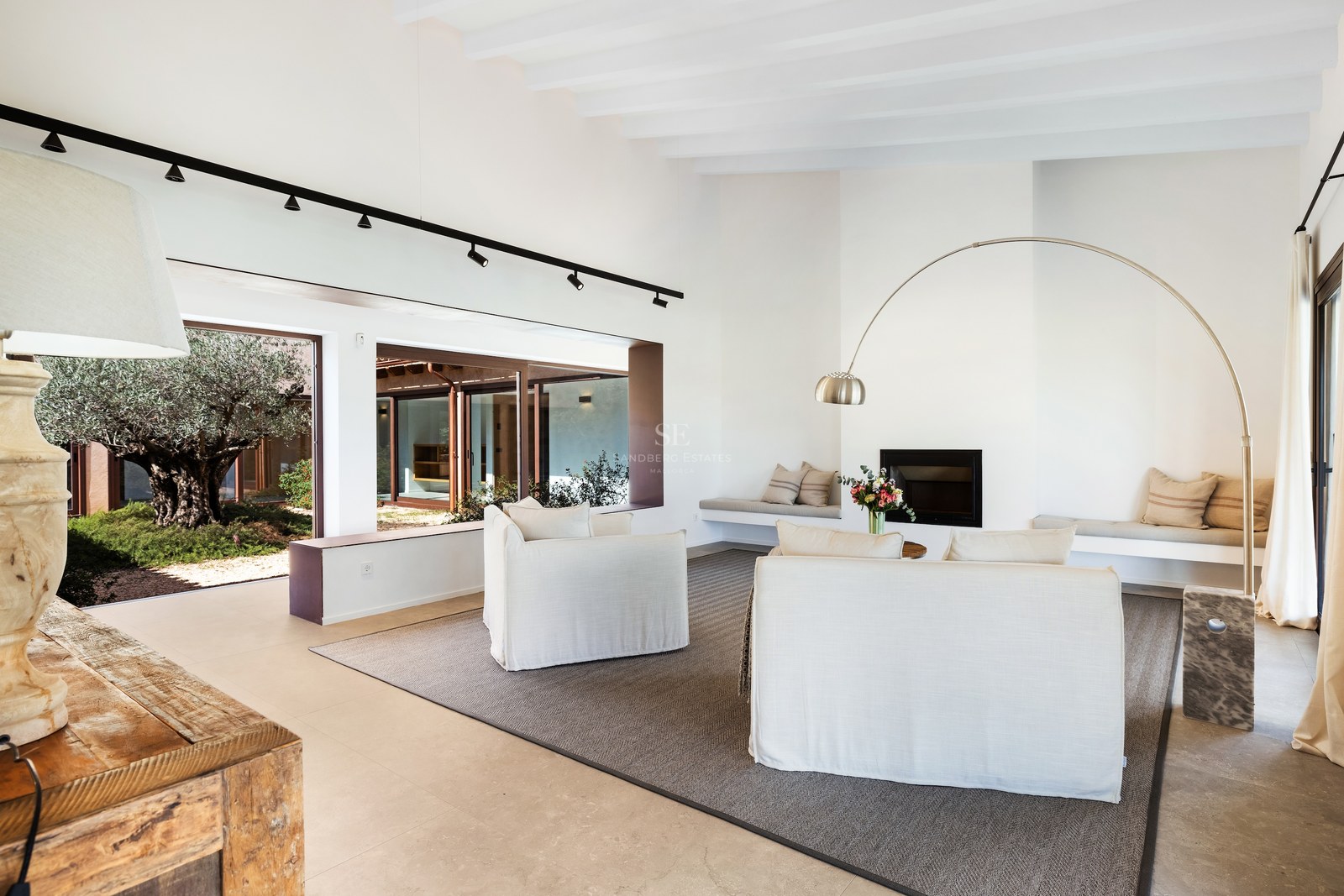 Bright living room with white sofas, an arched floor lamp, a fireplace, and views of an olive tree in a courtyard.