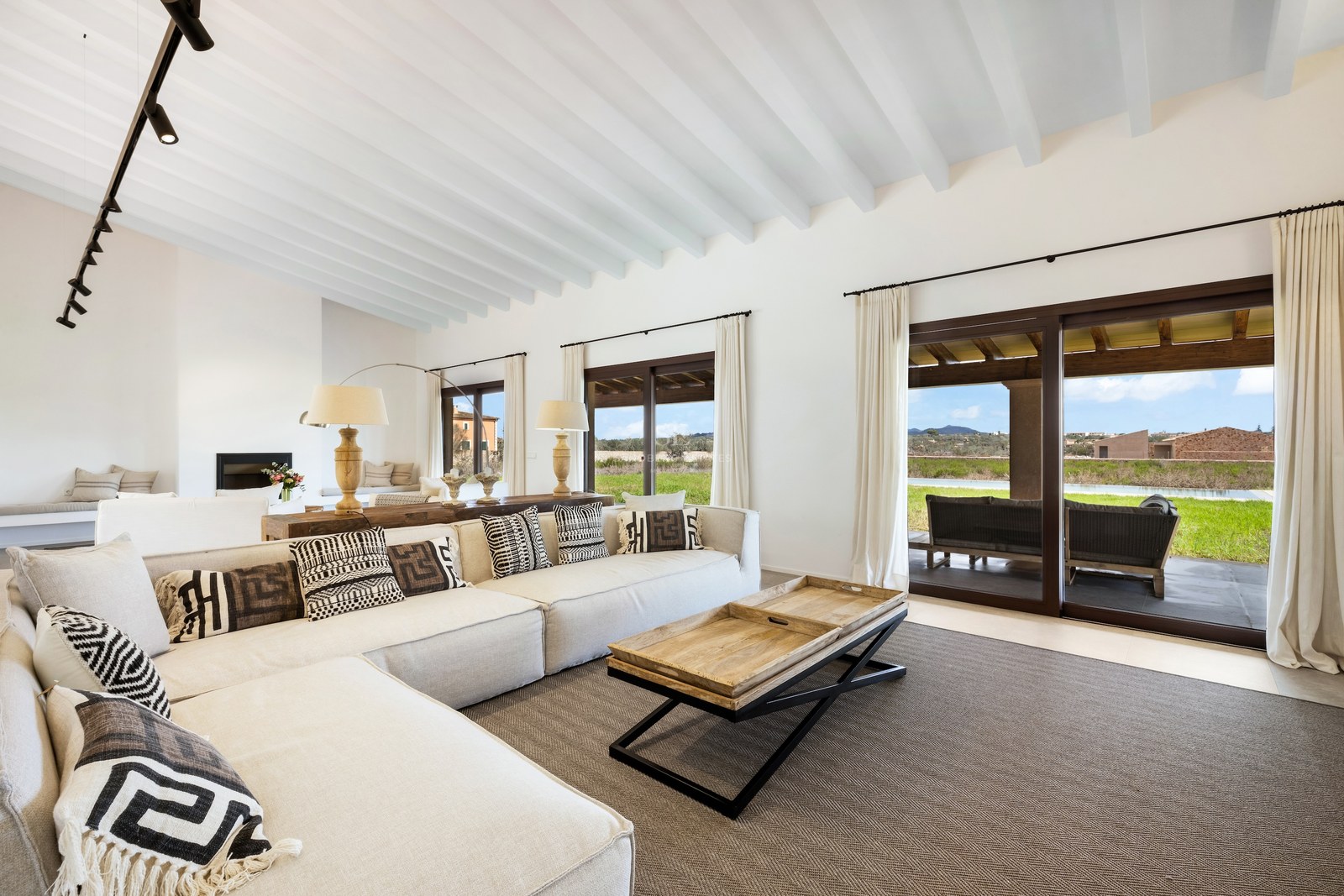 Bright living room with large L-shaped sofa, white wooden ceiling beams, and glass doors opening to a scenic terrace.