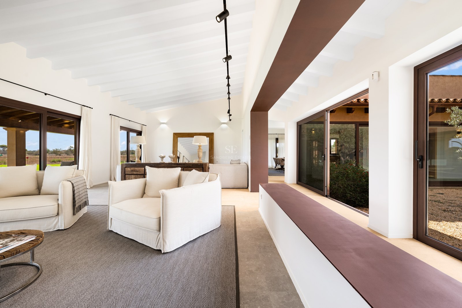 Spacious living room featuring white sofas, white beamed ceilings, and large glass doors opening to the outdoors.