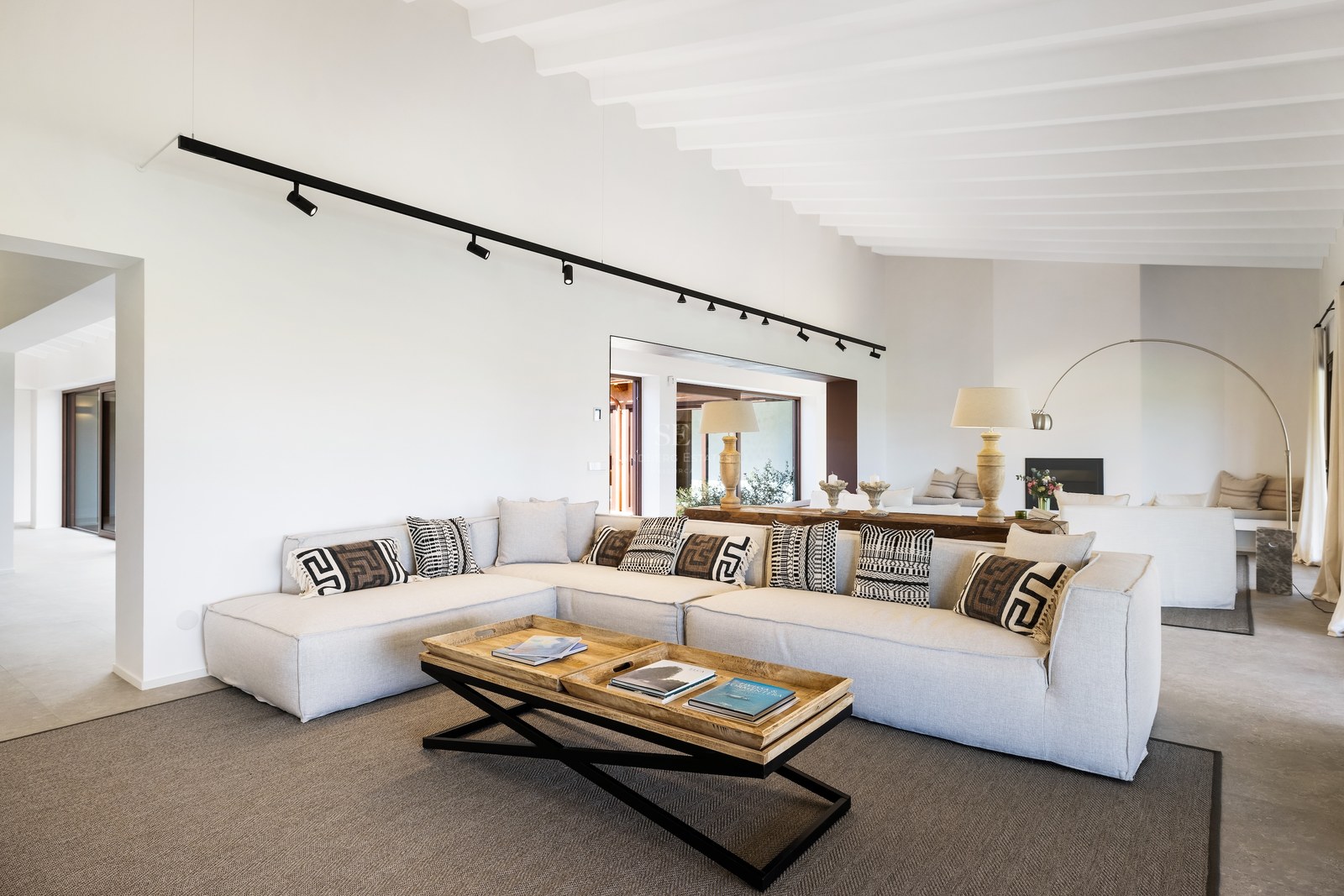 Spacious living room featuring a large grey sectional sofa, geometric cushions, and wooden coffee table under a white beamed ceiling.