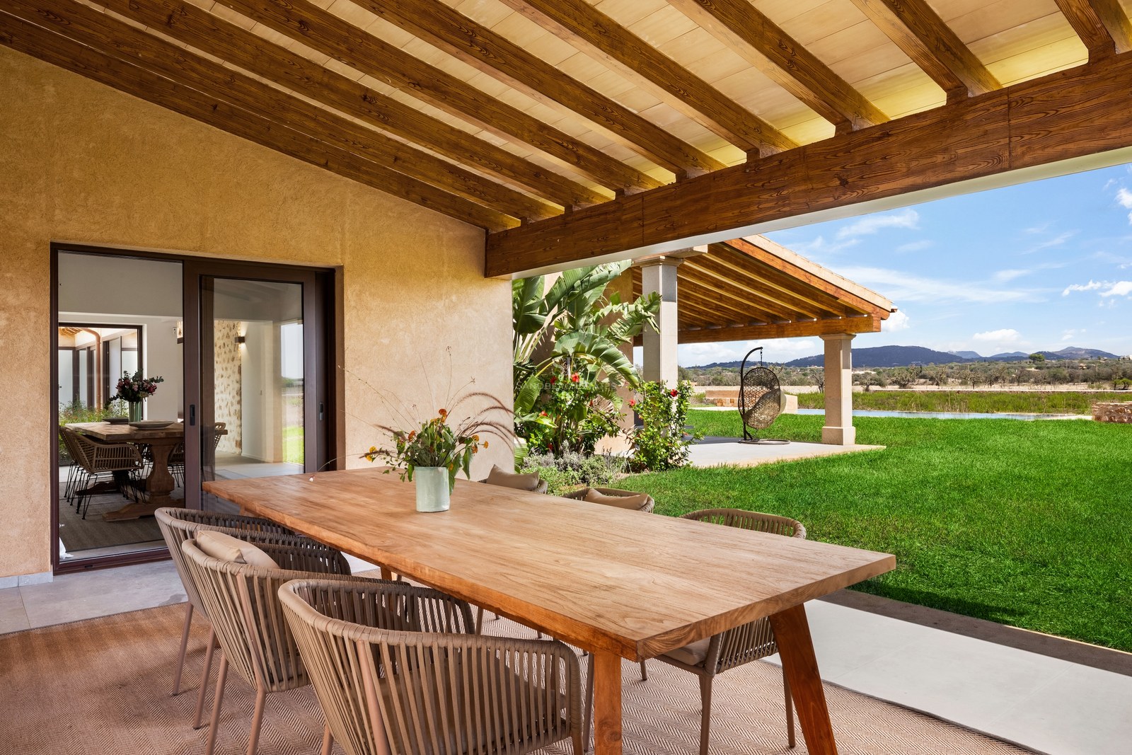 Covered terrace with a large wooden dining table and rope chairs overlooking a bright green lawn and distant hills.