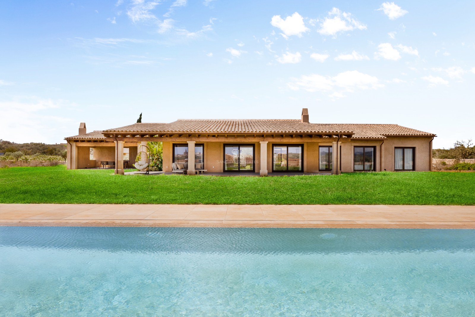 Large turquoise pool in front of a modern Mediterranean villa with a green lawn and terracotta roof under a blue sky.