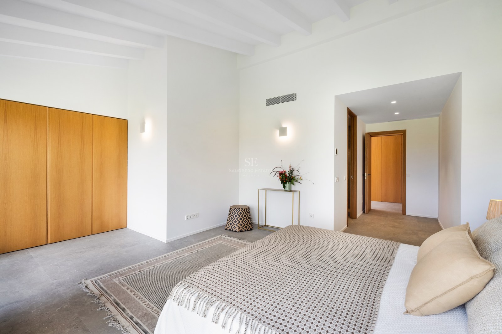 Bright bedroom with white beamed ceilings, wooden built-in wardrobes, large bed with beige pillows, and grey stone flooring.