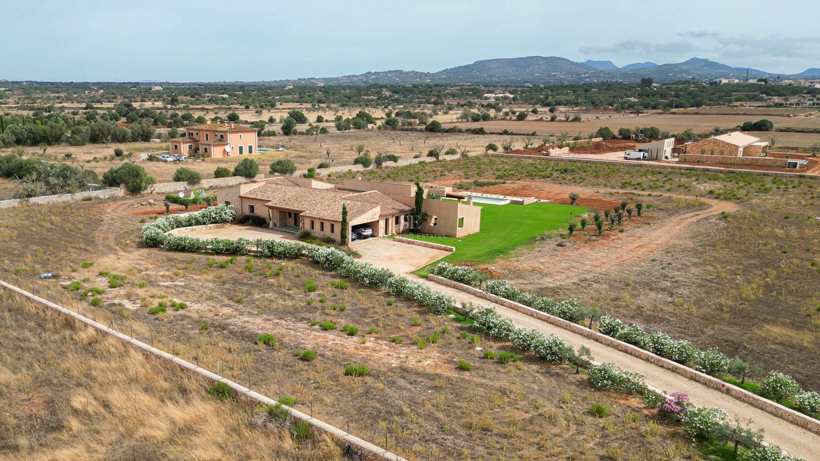 Aerial shot of a stone villa with a pool and flower-lined driveway in a Mediterranean countryside setting.