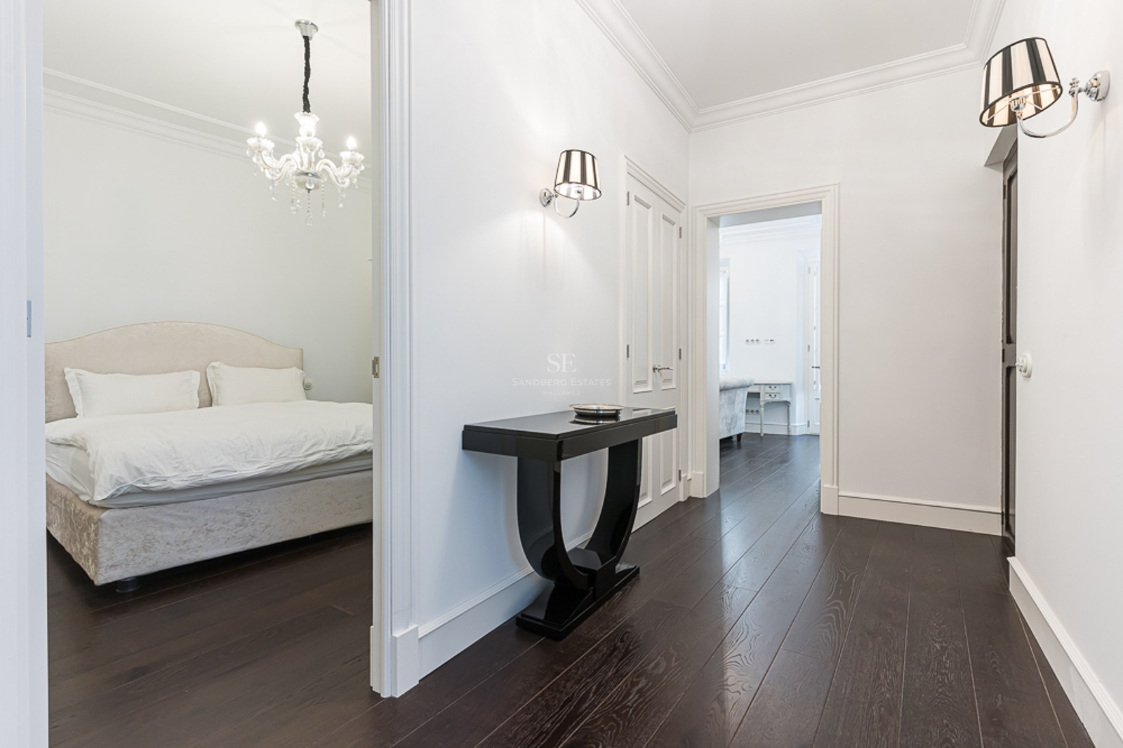 Bright hallway with dark wood floors, black console table, and view into a bedroom with a crystal chandelier.