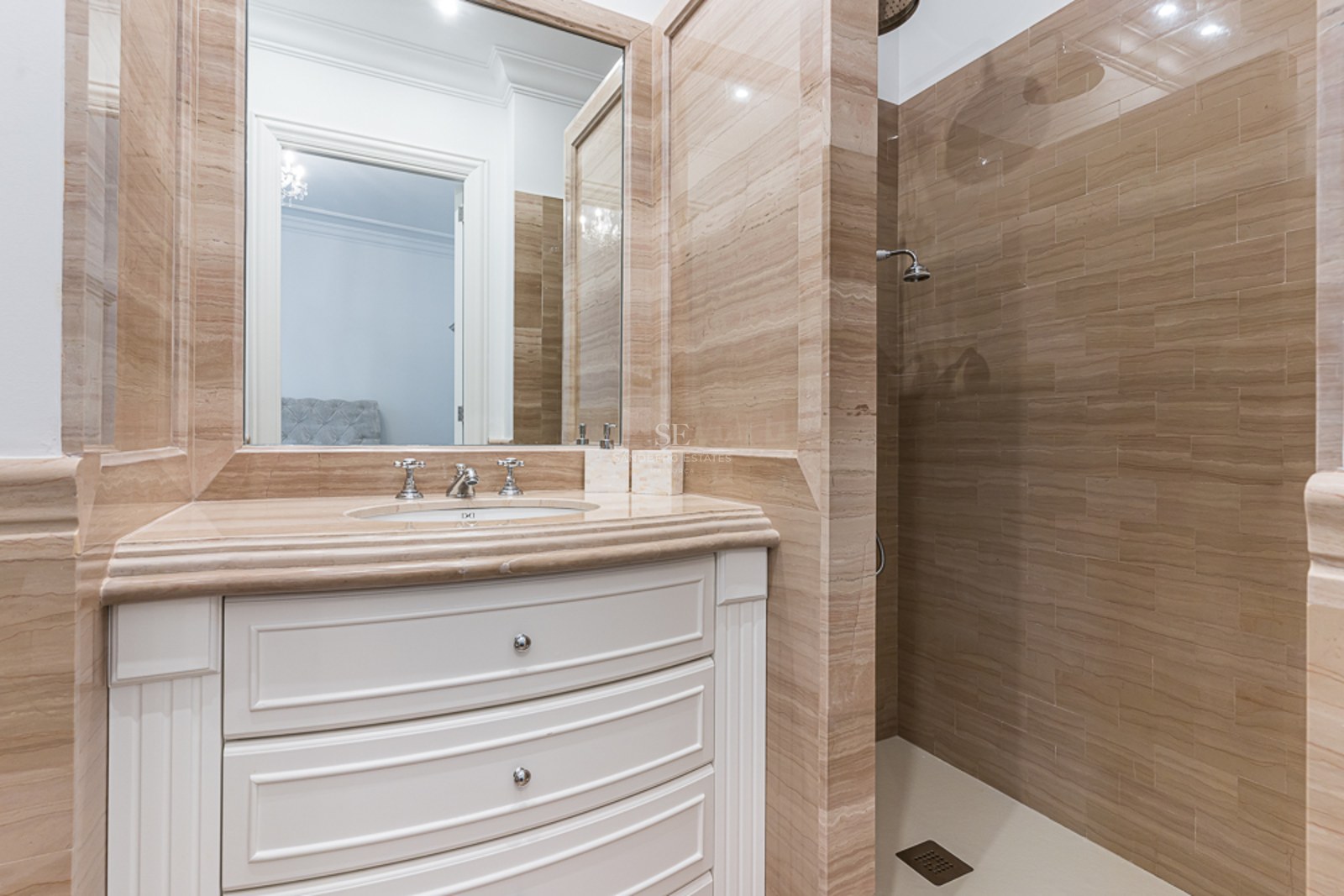 Luxury bathroom featuring beige travertine tile walls, a white curved vanity with marble top, and a walk-in shower.