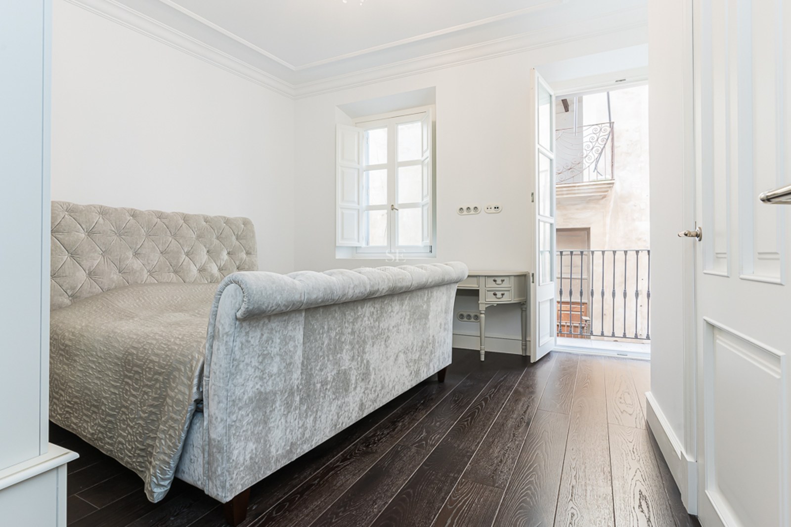 Bright bedroom featuring a grey velvet tufted bed, dark wood floors, and a balcony with wrought iron railing.