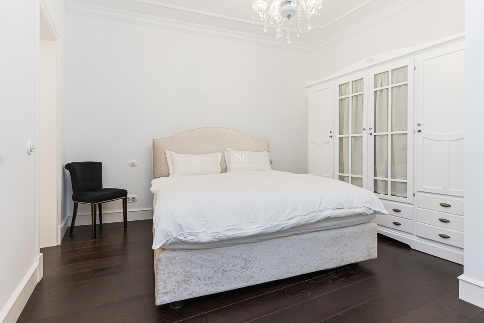Bright bedroom featuring a beige bed, large white wardrobe, black chair, and crystal chandelier on dark wood floors.