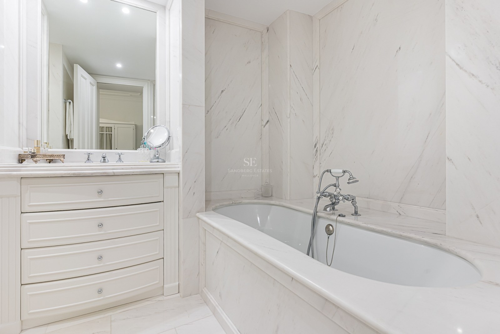 Bright master bathroom featuring white marble walls, a built-in tub with chrome faucets, and a white vanity with drawers.