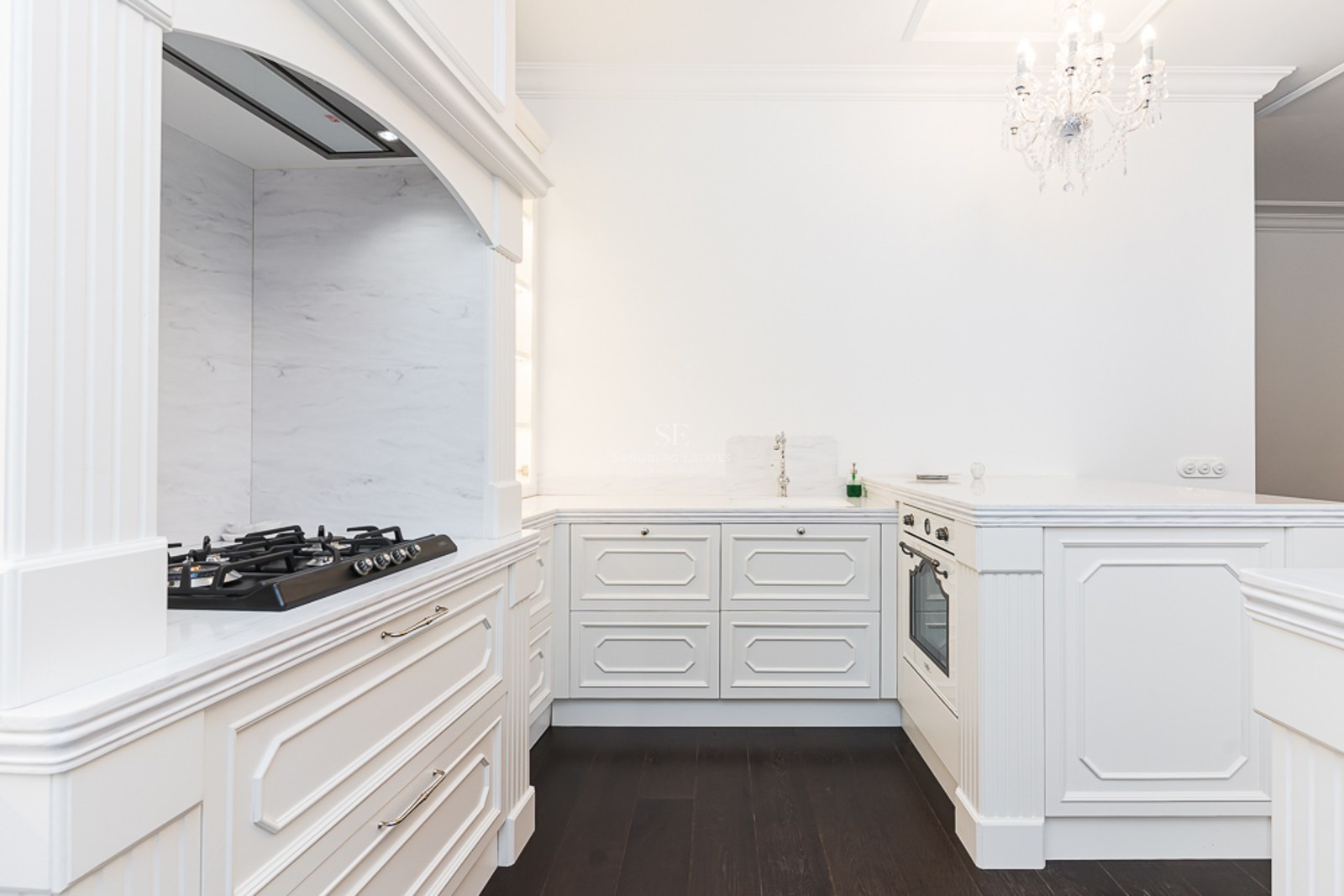 Elegant white kitchen with marble countertops, dark wood flooring, and a crystal chandelier.