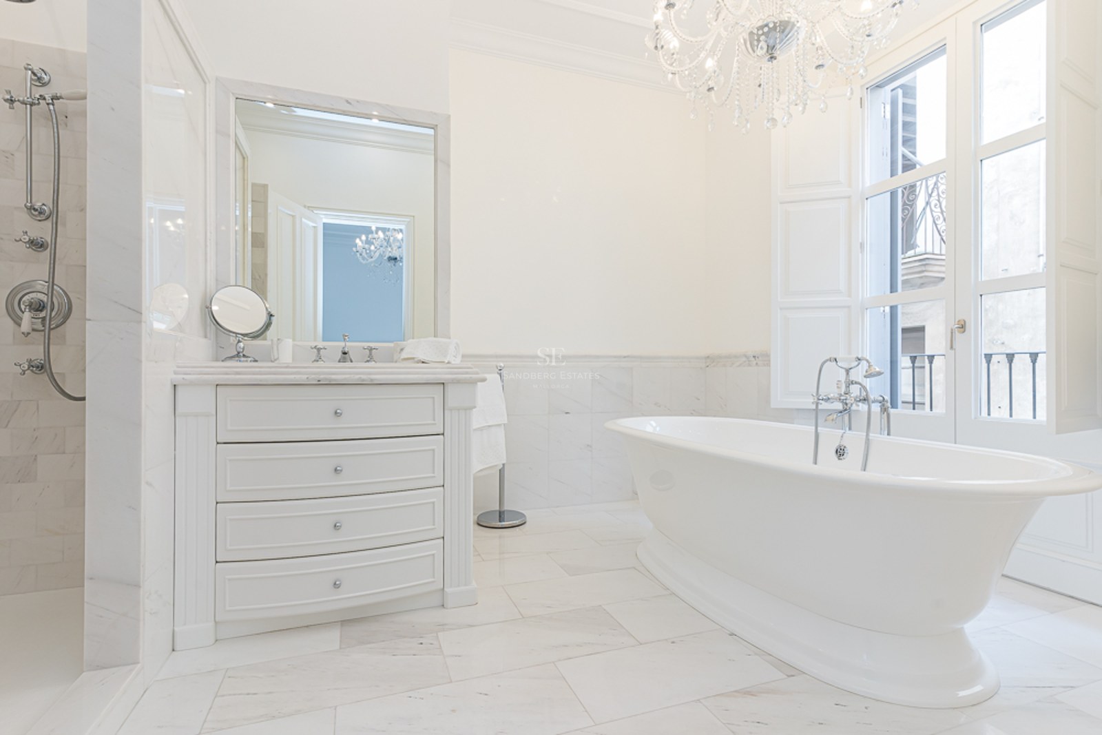 Luxury white marble bathroom featuring a freestanding tub, crystal chandelier, and large window with natural light.