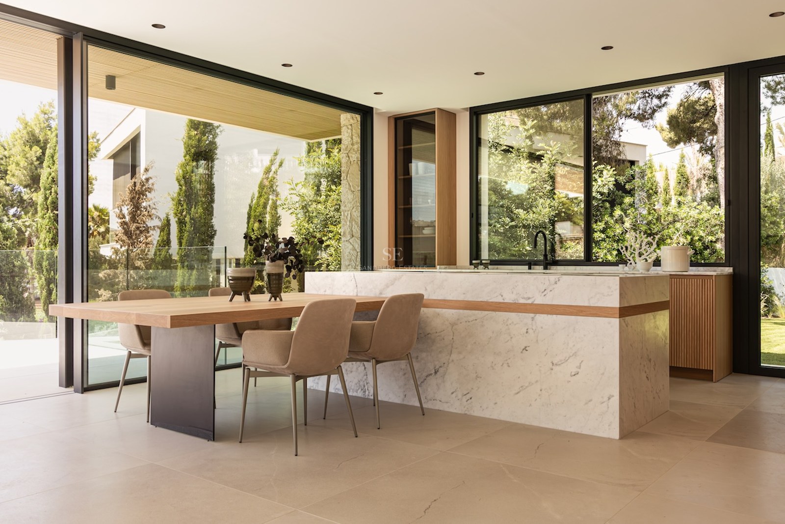 Salle à manger lumineuse avec table en bois, chaises beiges, îlot en marbre et grandes baies vitrées sur le jardin.