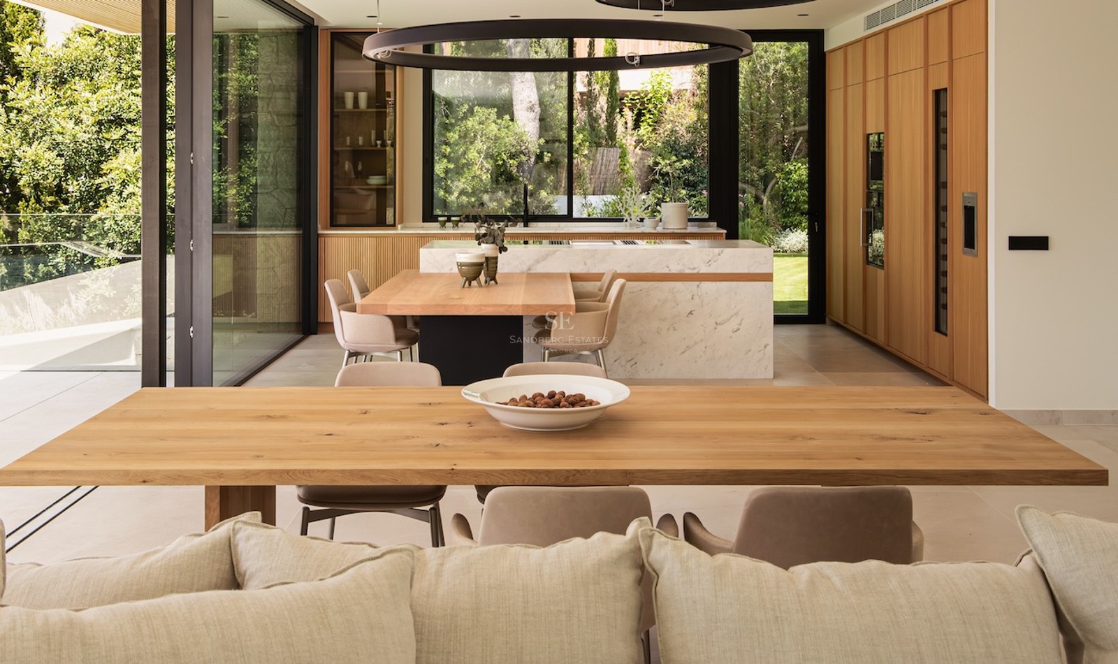 Salle à manger ouverte avec tables en bois, îlot en marbre et grandes vitres donnant sur un jardin verdoyant.