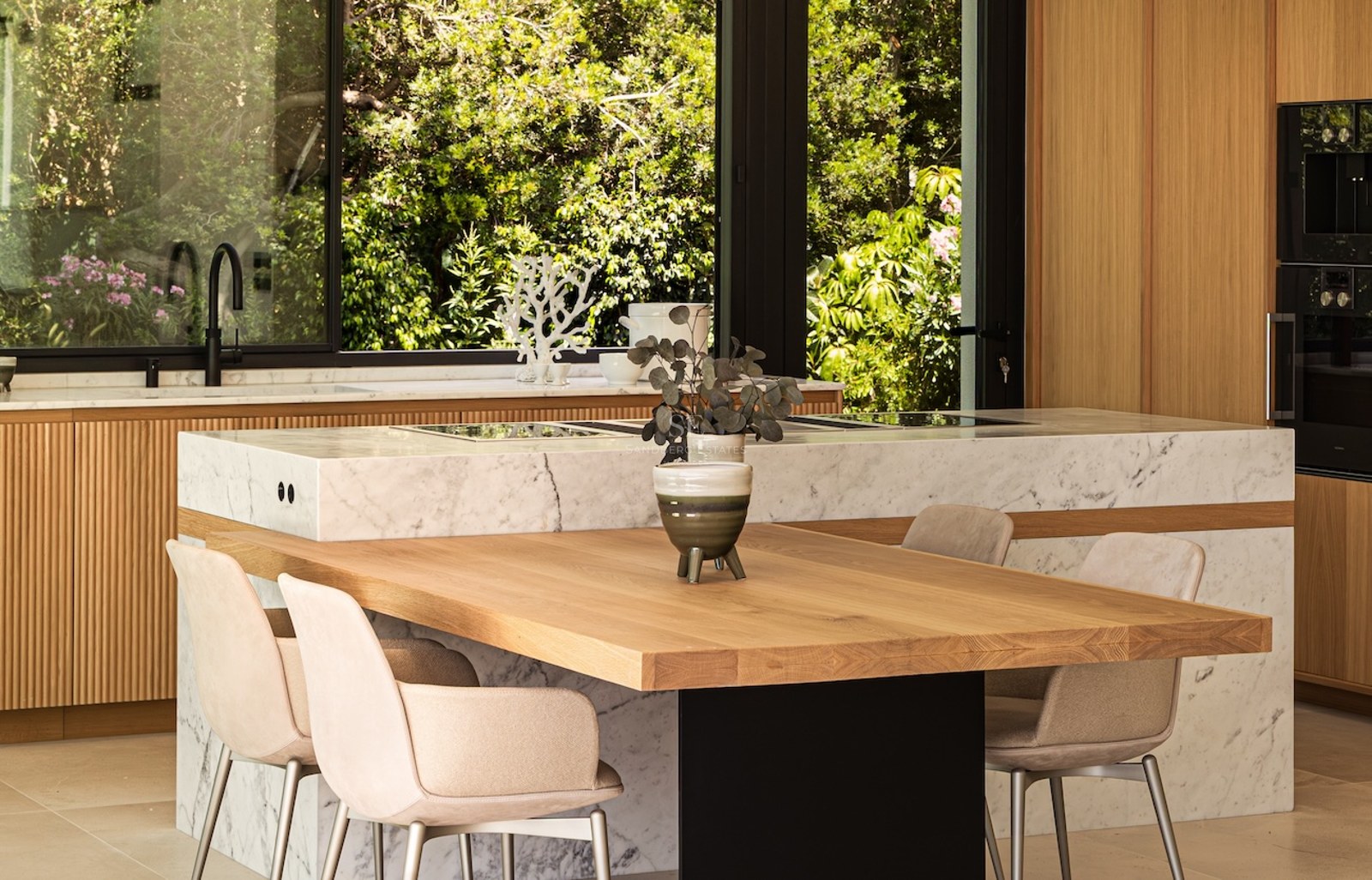 Modern kitchen featuring a white marble island with integrated wooden dining table and large windows showing lush gardens.