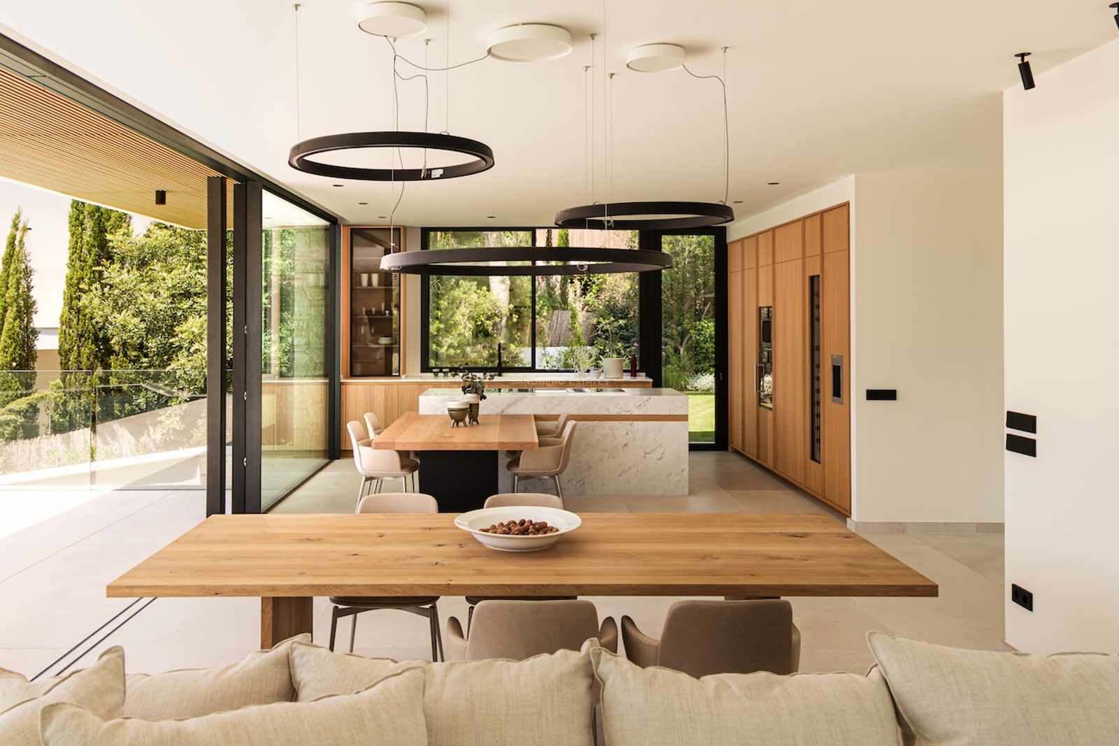 Salle à manger lumineuse avec table en bois, îlot en marbre, luminaires circulaires noirs et vue sur le jardin.