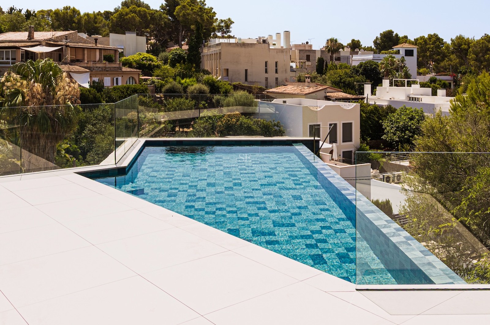Piscine à débordement aux carreaux bleus avec garde-corps en verre surplombant une ville méditerranéenne verdoyante.