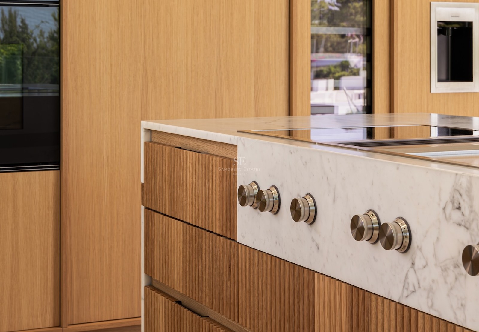 Close-up of a white marble kitchen island featuring brushed metal control knobs and fluted light wood cabinetry.