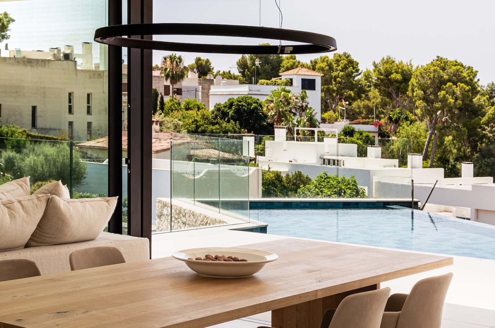Salle à manger lumineuse avec table en bois et vue sur une piscine et un paysage verdoyant à travers de grandes vitres.