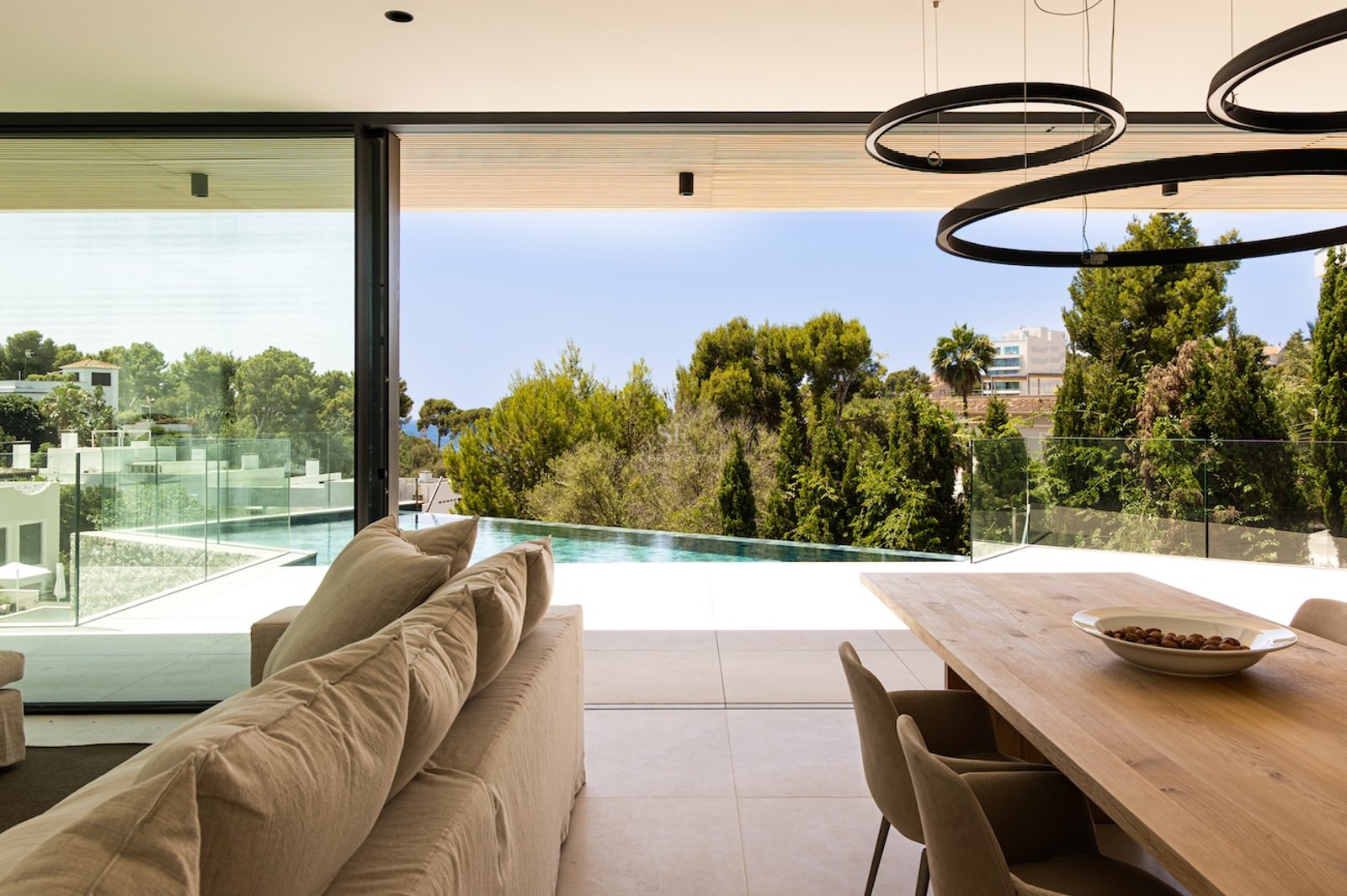 Salon lumineux avec canapé beige ouvrant sur une terrasse avec piscine à débordement et vue sur la nature.