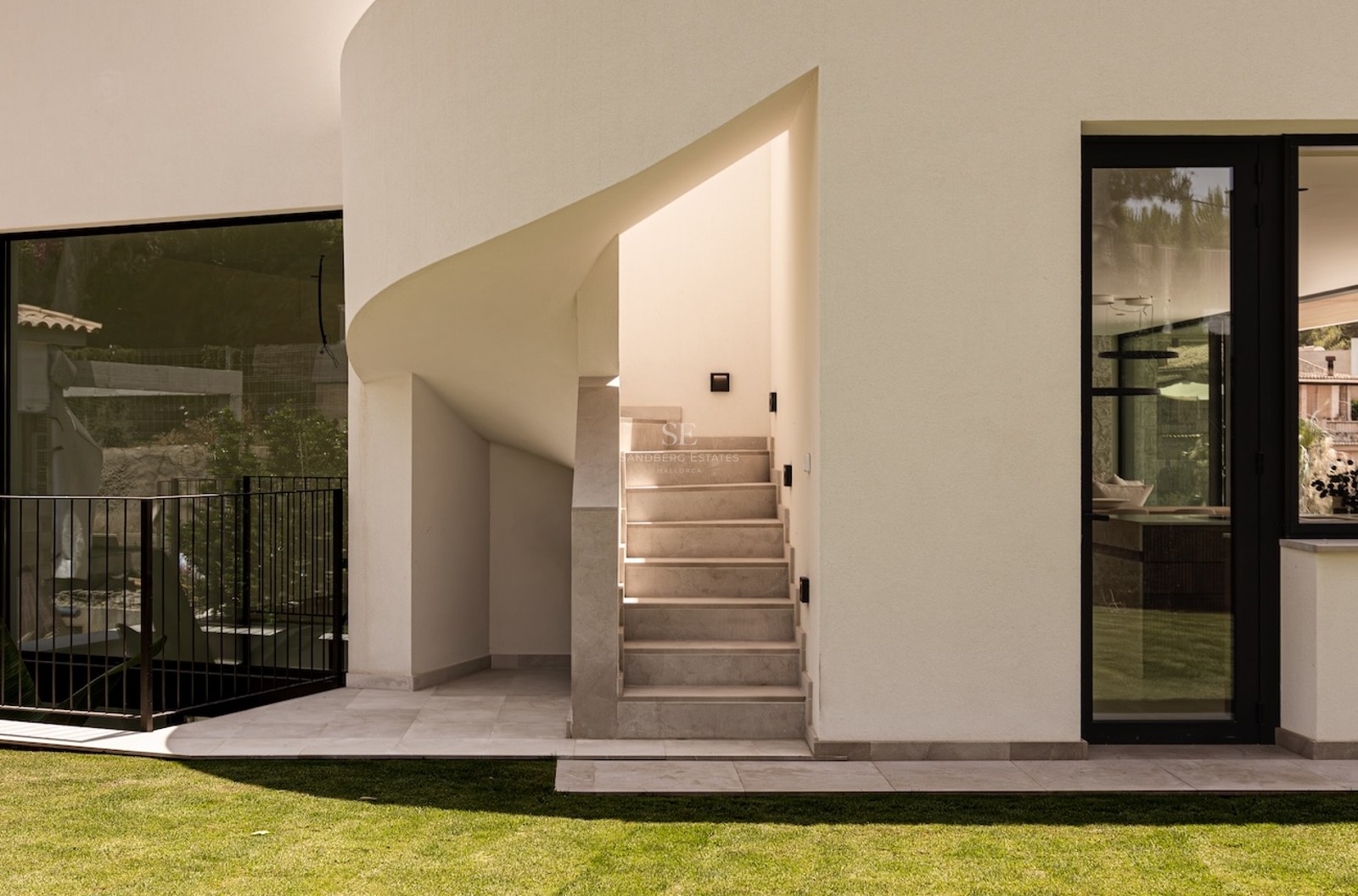 Exterior view of a modern white building featuring a stone staircase, a curved wall, and large black-framed windows.