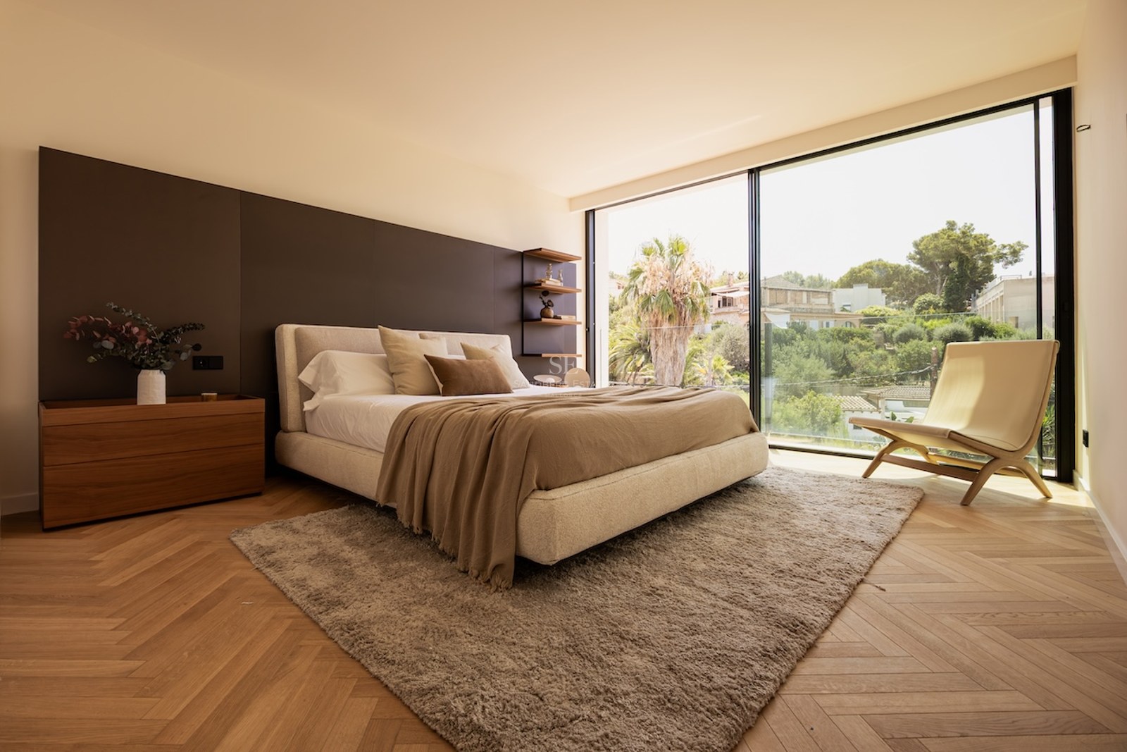Chambre moderne avec parquet en chevrons, grand lit, fauteuil design et baie vitrée ouvrant sur un jardin tropical.