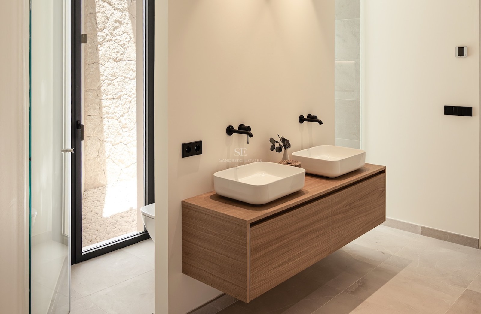 Salle de bain minimaliste avec double vasque sur meuble en bois, robinetterie noire et patio extérieur en pierre.
