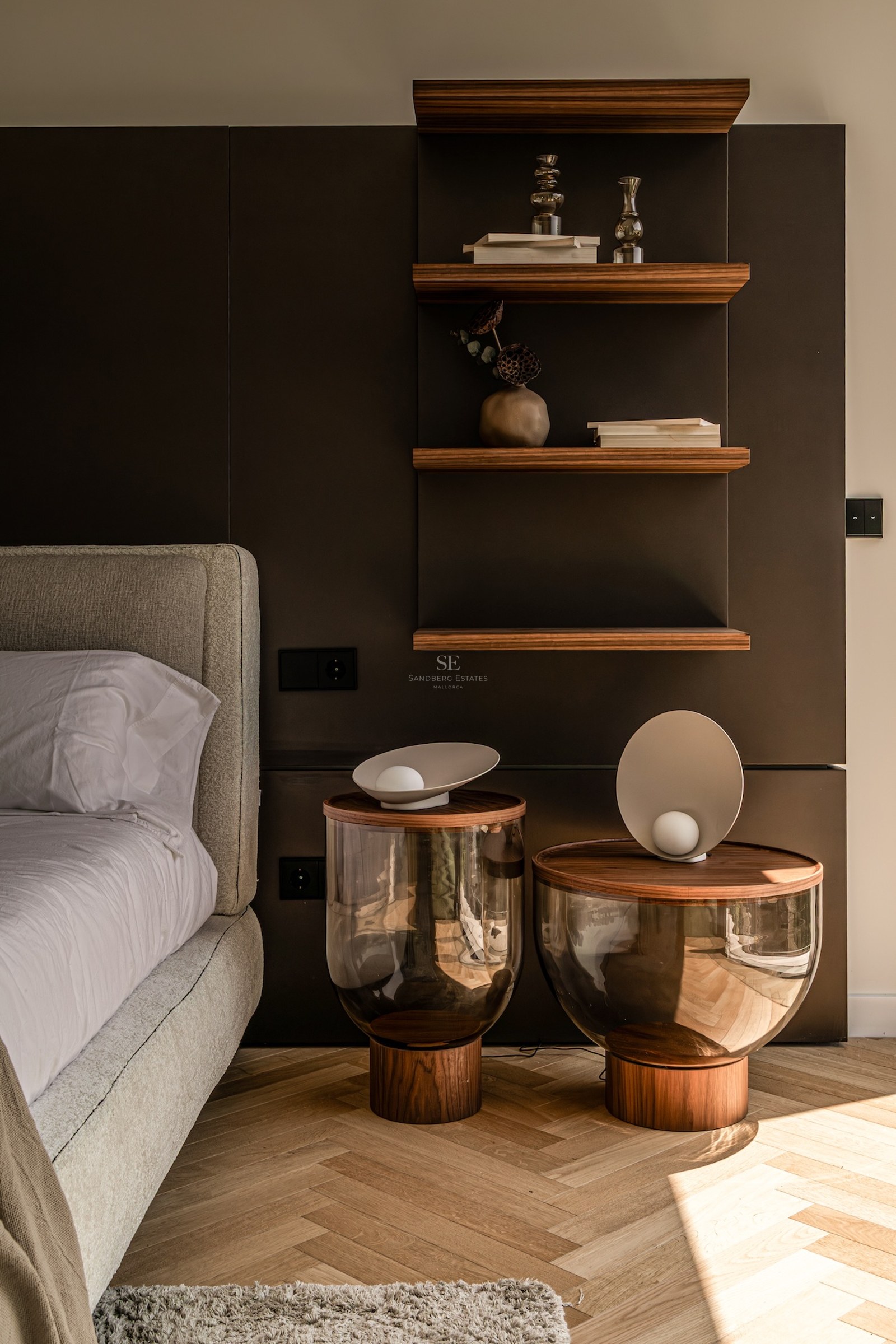 Modern bedroom corner featuring designer glass nightstands, floating walnut shelves, and herringbone oak flooring.