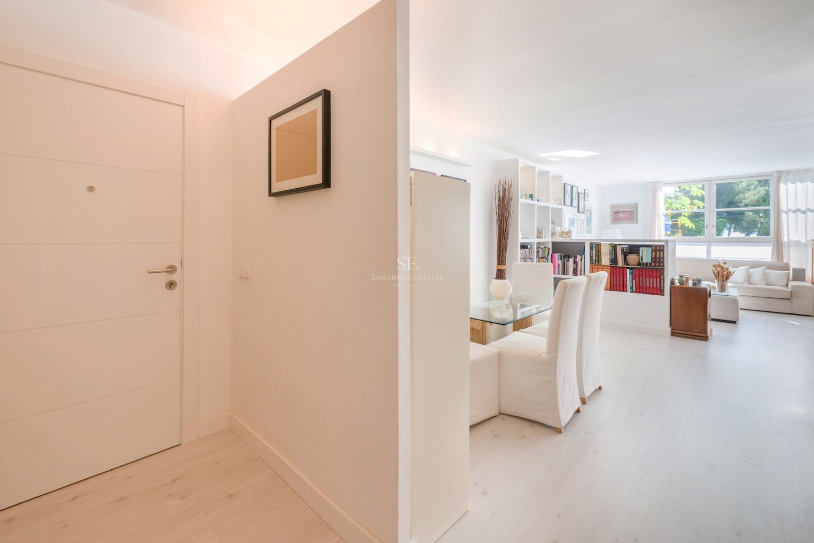 Une entrée blanche et lumineuse menant à un salon minimaliste avec du parquet en bois clair et de grandes fenêtres.