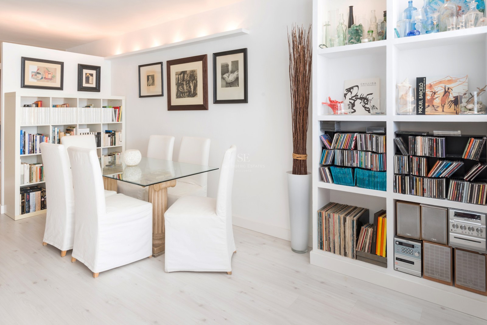 Une salle à manger lumineuse avec une table en verre, des chaises blanches et de grandes bibliothèques.