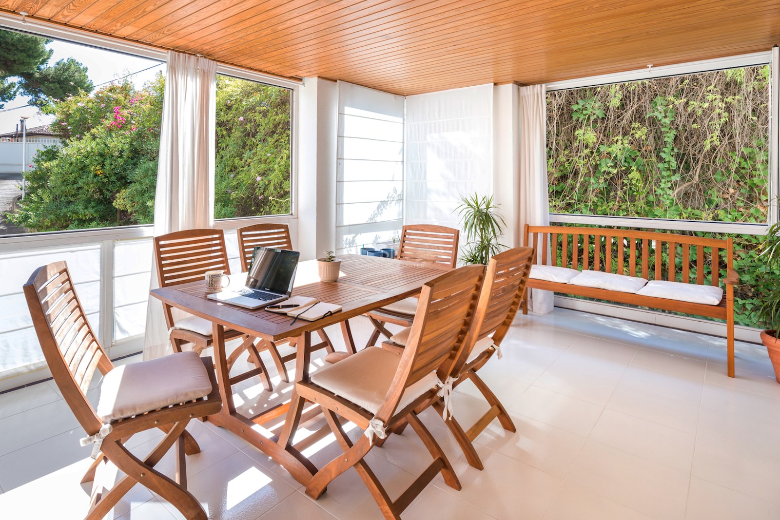 Une terrasse ensoleillée avec table à manger en bois, chaises et banc, donnant sur la verdure du jardin.