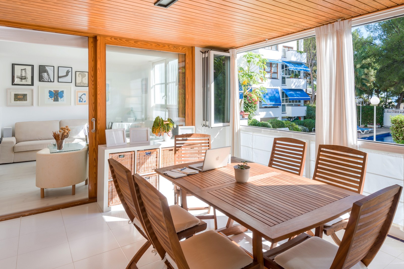 Terrasse fermée en bois avec table à manger et ordinateur, donnant sur une piscine et un jardin.