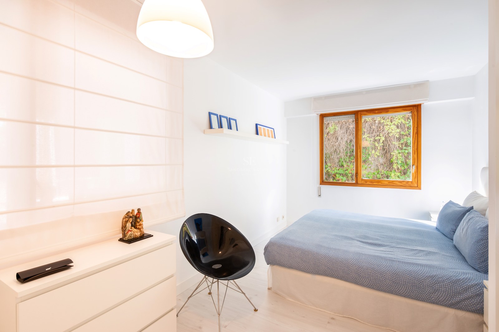 Chambre lumineuse avec grand lit, fenêtre en bois sur verdure, chaise design noire et commode blanche.