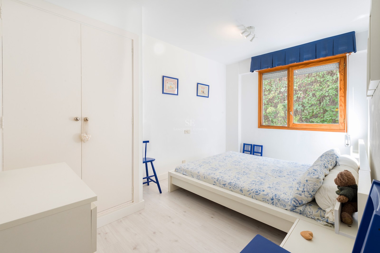 Chambre blanche et lumineuse avec une fenêtre à cadre en bois, un placard intégré et une literie florale bleue.