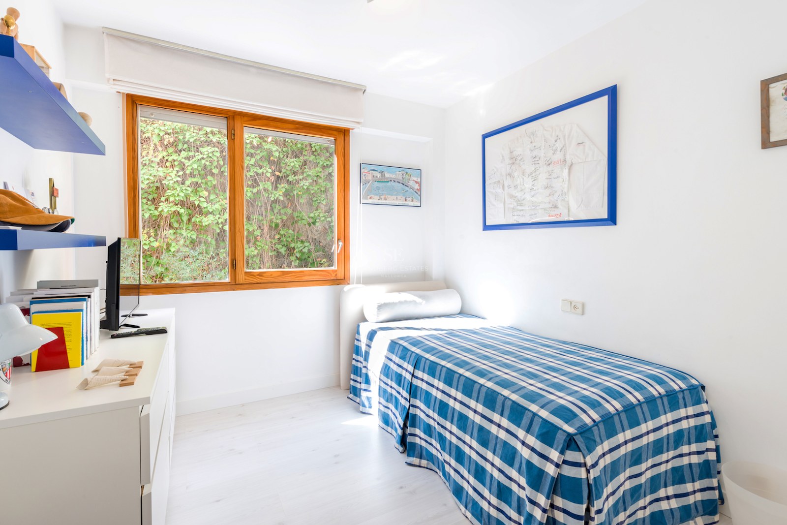 Chambre simple lumineuse avec un lit à carreaux bleus, un bureau blanc et une grande fenêtre en bois.