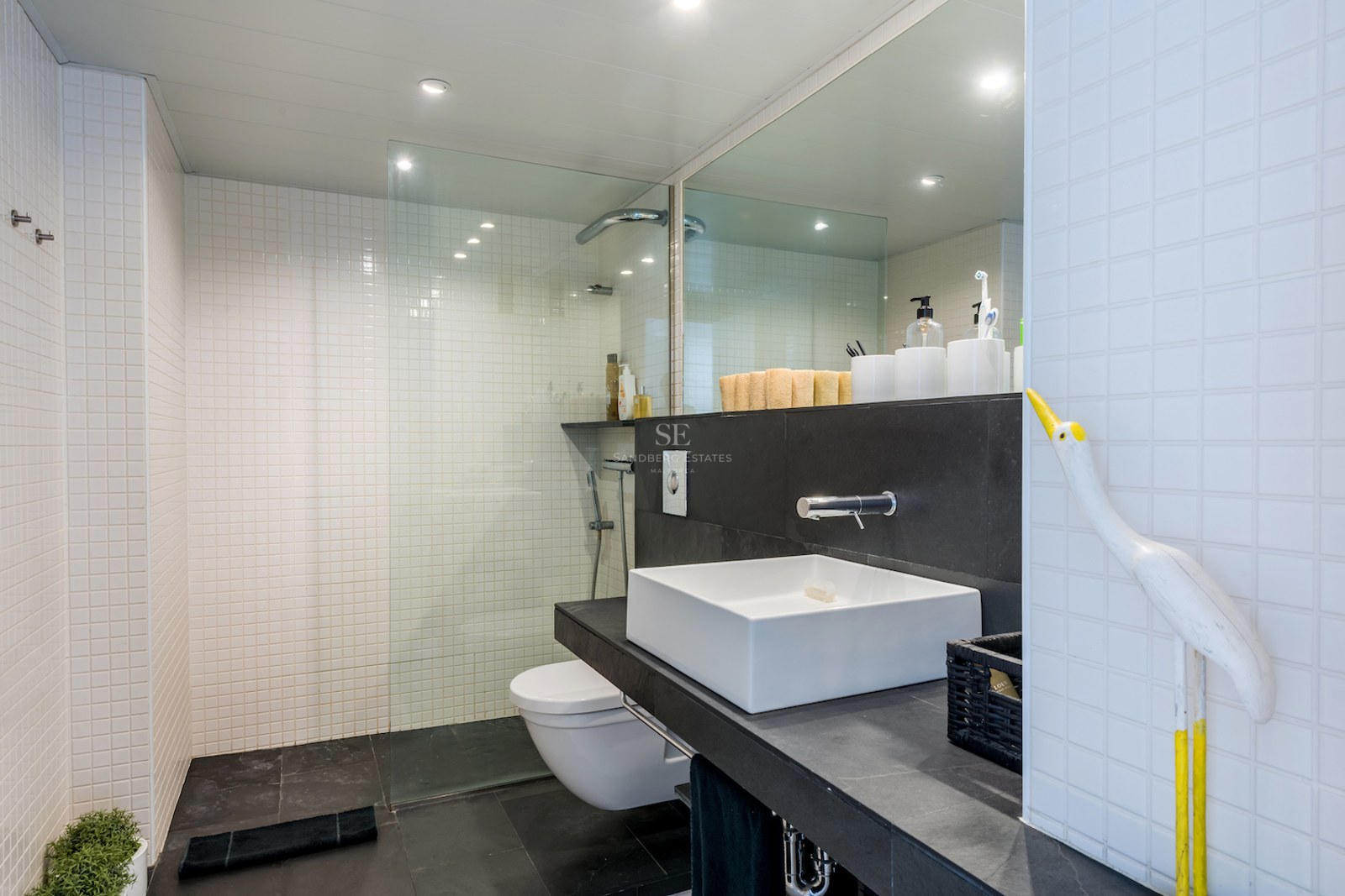 Salle de bain moderne avec carrelage blanc, sol noir, douche en verre et vasque carrée.