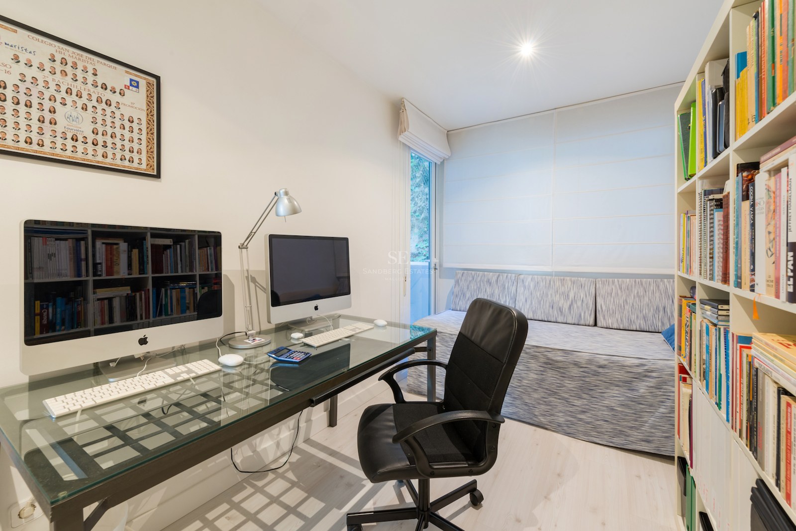 Un bureau lumineux avec un bureau en verre, deux ordinateurs, une chaise ergonomique noire et de grandes étagères.