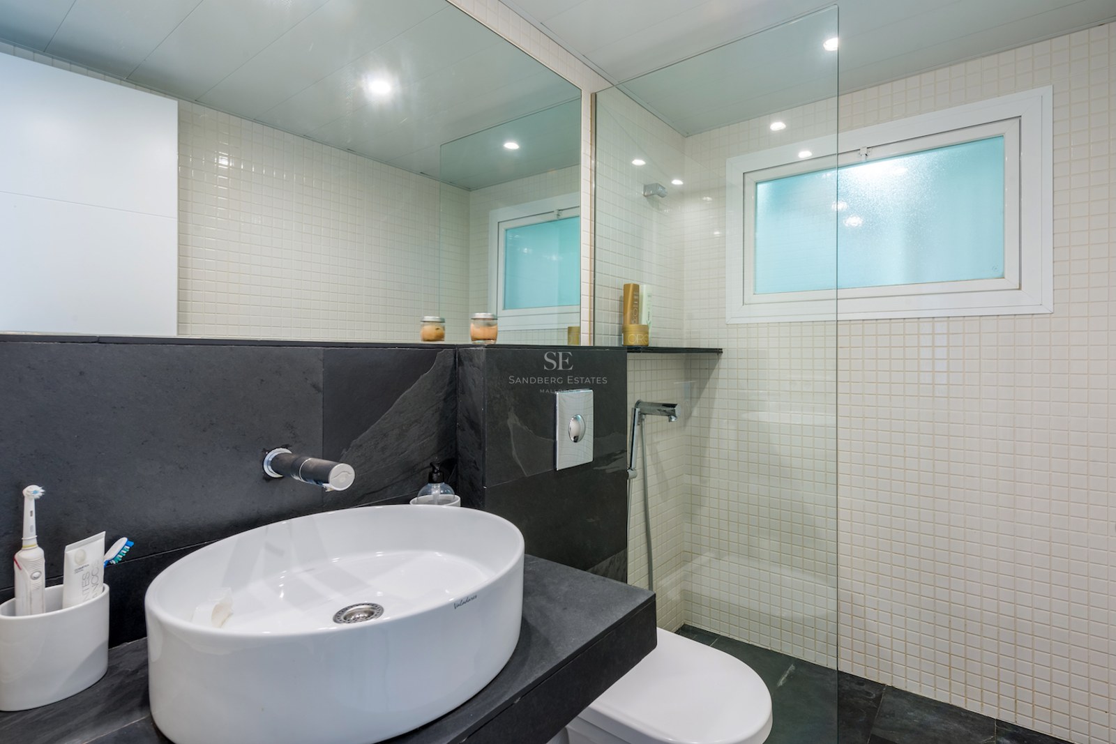 Salle de bain moderne avec vasque ovale blanche sur plan en ardoise foncée, mosaïque blanche et paroi de douche en verre.