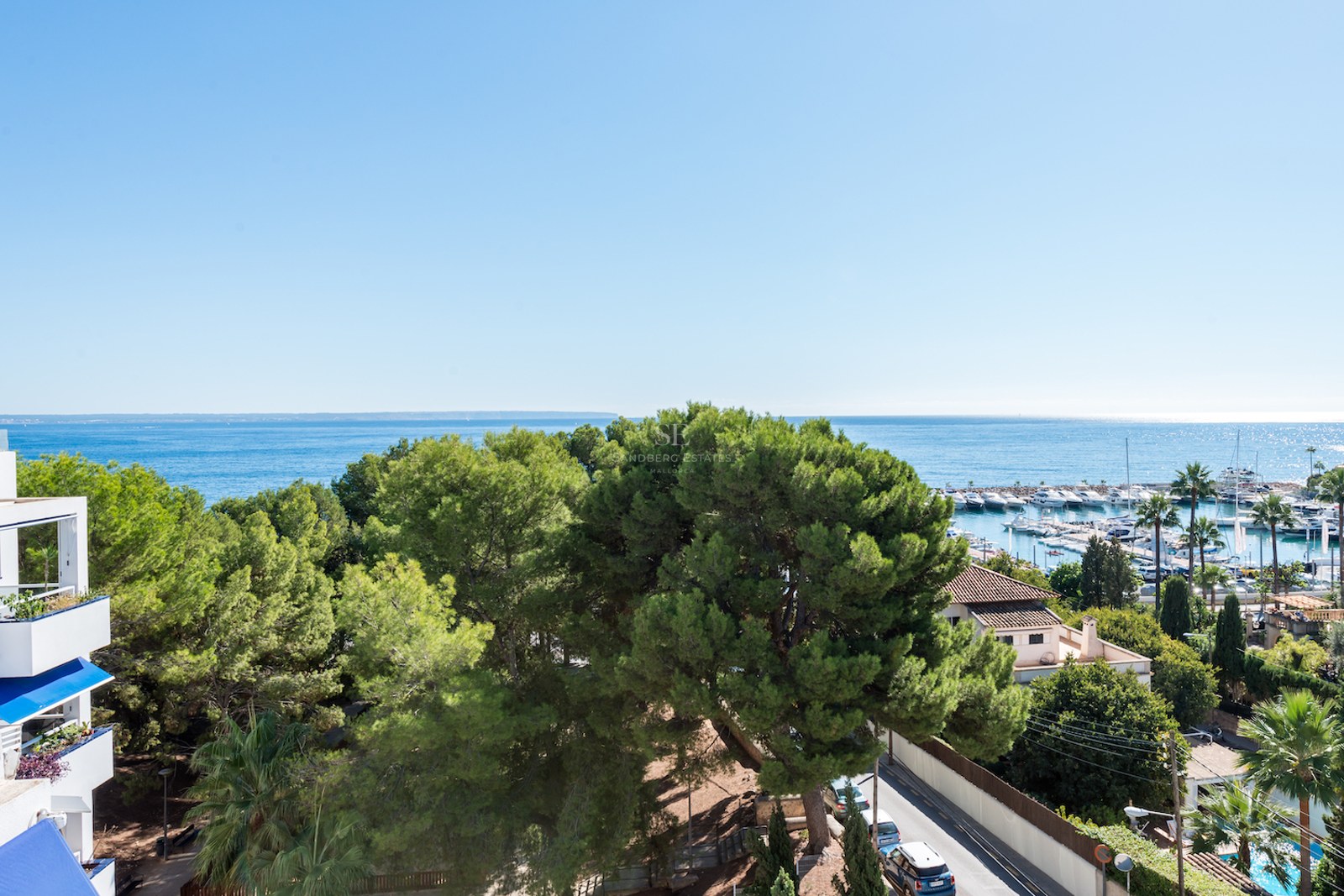 Vue en plongée sur la mer Méditerranée bleue, une marina avec des bateaux et des pins verts sous un ciel dégagé.