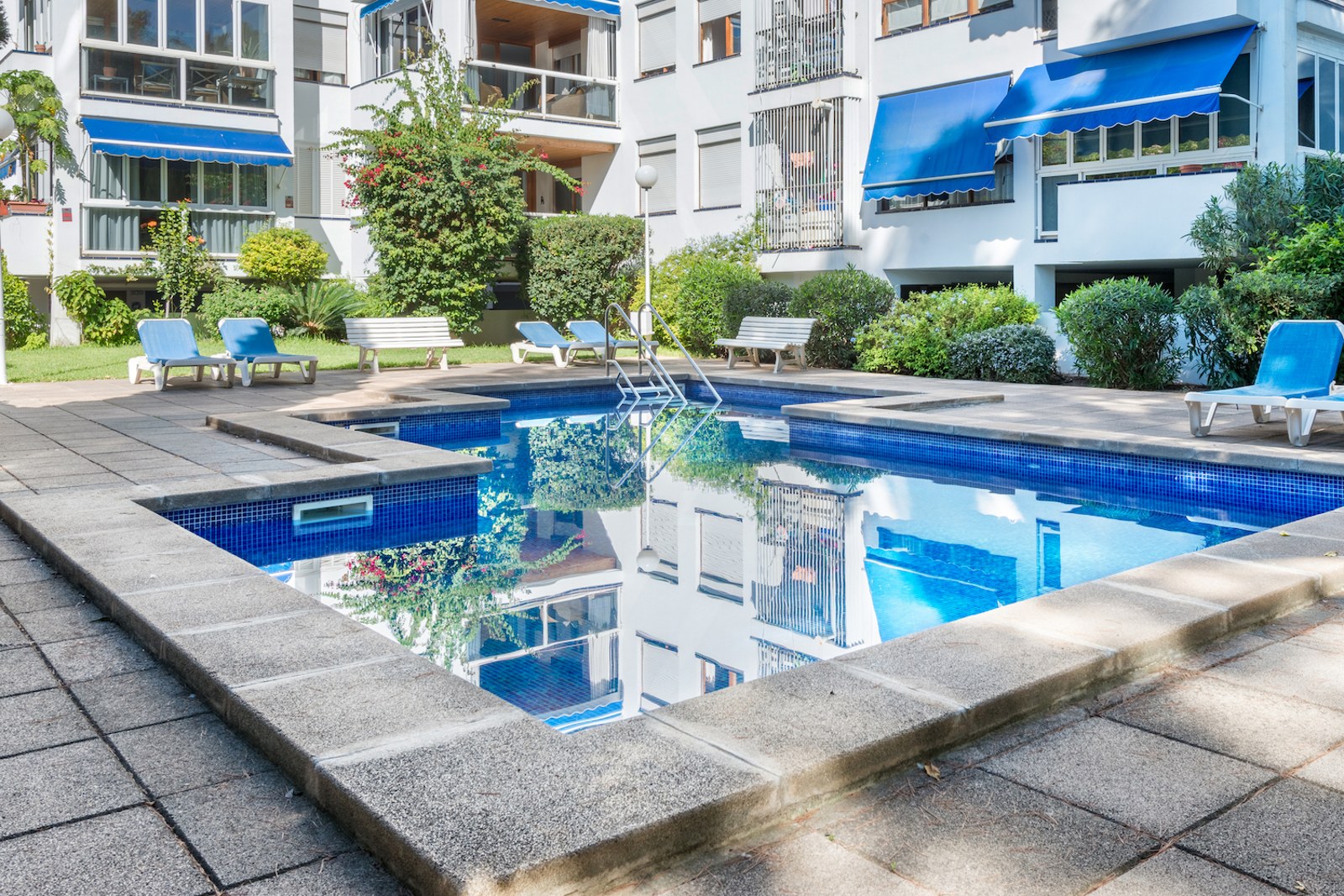 Piscine rectangulaire bleue entourée d'une terrasse en pierre avec des transats devant un immeuble blanc.