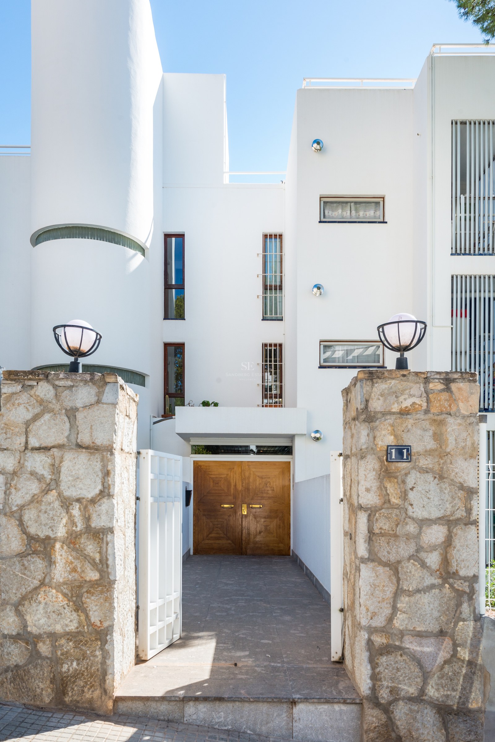 High-contrast view of a white villa entrance featuring natural stone pillars and solid wooden doors in bright daylight.