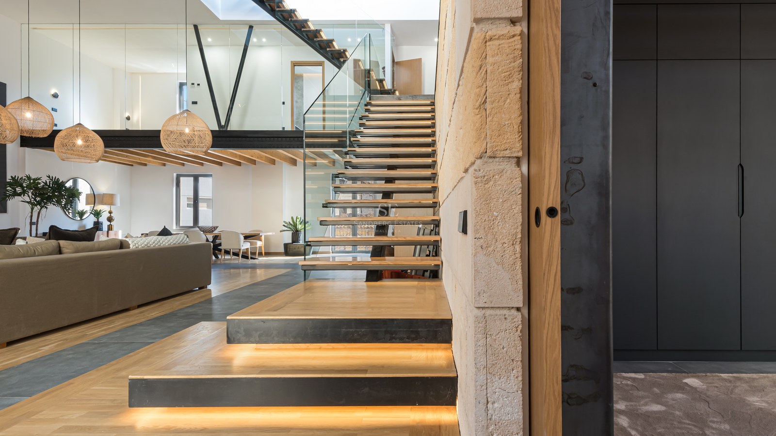 Modern open-plan living room featuring a wooden floating staircase with LED lights, glass railings, and a mezzanine.